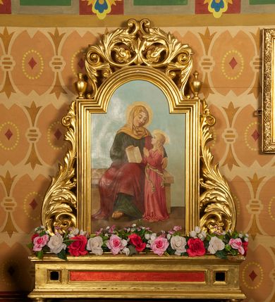 Zdjęcie nr 1: Feretron z obrazem Święta Anna nauczająca Marię. Podstawa w kształcie leżącego prostopadłościanu wspartego na kulistych nóżkach, profilowana, z w otworami na drążki, złocona, z czerwoną płyciną pośrodku. Obraz otoczony złoconą, profilowaną ramą w formie stojącego prostokąta zamkniętego od góry łukiem półkolistym nadwieszonym. Rama flankowana liśćmi akantu tworzącymi spływy wolutowe, ze zwieńczeniem utworzonym z liści akantu zwijanych w symetryczne esownice. Na uskokach ramy niewielkie wazony.
W polu obraz przedstawiający Annę z Marią. W centrum kompozycji Anna siedząca na ławie, zwrócona w trzech czwartych w lewo, z głową i rękoma skierowanymi w prawo, w stronę Marii, prawą ręką przytrzymuje otwartą księgę, opartą na kolanie, a lewą obejmuje córkę. Twarz ma podłużną, o dużych oczach, uniesionych brwiach, prostym nosie i małych ustach. Na głowie ma jasnożółty welon, jest ubrana w długą, ciemnozieloną suknię i czerwony płaszcz okrywający jej nogi. Spod szat wystaje jej prawa stopa w sandale, wsparta na stopniu. Maria stoi przy lewym boku matki, ukazana jest z profilu, z głową skierowaną w górę i dłońmi złożonymi w geście modlitwy. Ma pociągłą twarz o delikatnych rysach; okoloną długimi, jasnymi i falowanymi włosami. Jest ubrana w długą, różową suknię, przewiązaną w pasie wstążką, której końce skręcają się i rozwiewają do boku. Obie postacie mają wokół głów świetliste nimby.Tło sceny jednolite, jasnoniebieskie, w dolnej partii obrazu widoczna jasnobrązowa posadzka.
