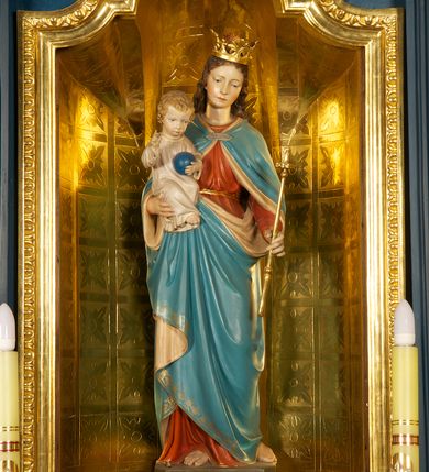Zdjęcie nr 1: Figura Matki Boskiej z Dzieciątkiem ustawiona na niskiej, ciemnobrązowej podstawie. Maria ukazana frontalnie, w wykroku, z głową lekko przechyloną w lewo; prawą ręką podtrzymuje Dzieciątko, w lewej trzyma złote berło. Twarz ma owalną, o młodzieńczych i regularnych rysach; oczy ma skierowane w dół, prosty, długi nos i wąskie usta. Jasnobrązowe, falowane włosy spływają jej na plecy. Na głowie ma złoconą, otwartą koronę. Jest ubrana w czerwoną suknię przewiązaną w pasie złoconym sznurem oraz niebieski płaszcz z beżową podszewką, spięty na piersi klamrą, okrywający ramiona, nogi i podtrzymywany z przodu. Dzieciątko w pozycji siedzącej, frontalnie, ze skrzyżowanymi nogami, prawą rękę unosi w geście błogosławieństwa, w lewej trzyma niebieską kulę. Ma owalną twarz, małe oczy, wąski nos i niewielkie usta, na policzkach rumieńce, a na głowie krótkie, jasne, kręcone włosy. Jest ubrane w jasną, długą tunikę.