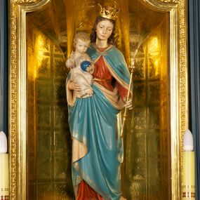 Zdjęcie nr 1: Figura Matki Boskiej z Dzieciątkiem ustawiona na niskiej, ciemnobrązowej podstawie. Maria ukazana frontalnie, w wykroku, z głową lekko przechyloną w lewo; prawą ręką podtrzymuje Dzieciątko, w lewej trzyma złote berło. Twarz ma owalną, o młodzieńczych i regularnych rysach; oczy ma skierowane w dół, prosty, długi nos i wąskie usta. Jasnobrązowe, falowane włosy spływają jej na plecy. Na głowie ma złoconą, otwartą koronę. Jest ubrana w czerwoną suknię przewiązaną w pasie złoconym sznurem oraz niebieski płaszcz z beżową podszewką, spięty na piersi klamrą, okrywający ramiona, nogi i podtrzymywany z przodu. Dzieciątko w pozycji siedzącej, frontalnie, ze skrzyżowanymi nogami, prawą rękę unosi w geście błogosławieństwa, w lewej trzyma niebieską kulę. Ma owalną twarz, małe oczy, wąski nos i niewielkie usta, na policzkach rumieńce, a na głowie krótkie, jasne, kręcone włosy. Jest ubrane w jasną, długą tunikę.