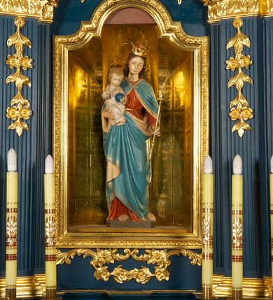 Zdjęcie nr 2: Figura Matki Boskiej z Dzieciątkiem ustawiona na niskiej, ciemnobrązowej podstawie. Maria ukazana frontalnie, w wykroku, z głową lekko przechyloną w lewo; prawą ręką podtrzymuje Dzieciątko, w lewej trzyma złote berło. Twarz ma owalną, o młodzieńczych i regularnych rysach; oczy ma skierowane w dół, prosty, długi nos i wąskie usta. Jasnobrązowe, falowane włosy spływają jej na plecy. Na głowie ma złoconą, otwartą koronę. Jest ubrana w czerwoną suknię przewiązaną w pasie złoconym sznurem oraz niebieski płaszcz z beżową podszewką, spięty na piersi klamrą, okrywający ramiona, nogi i podtrzymywany z przodu. Dzieciątko w pozycji siedzącej, frontalnie, ze skrzyżowanymi nogami, prawą rękę unosi w geście błogosławieństwa, w lewej trzyma niebieską kulę. Ma owalną twarz, małe oczy, wąski nos i niewielkie usta, na policzkach rumieńce, a na głowie krótkie, jasne, kręcone włosy. Jest ubrane w jasną, długą tunikę.