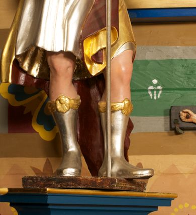 Zdjęcie nr 6: Figura ustawiona na niskiej, prostokątnej podstawie. Ukazana frontalnie, w wykroku lewej nogi, w lekkim odchyleniu ciała, z uniesioną głową skierowaną w lewo. Obie ręce ma wyciągnięte przed siebie, w prawej trzyma włócznię. Twarz ma szeroką, z niskim czołem i podkreślonym podbródkiem, o małych, migdałowatych oczach, niewielkim nosie i ustach. Włosy rzeźbione w lekko falowane pasma, ściśle przylegają do głowy i opadają na kark. Postać ma na głowie koronę otwartą. Ubrana jest w srebrzoną tunikę, sięgającą kolan, z podwiniętymi rękawami, złoconą zbroję tortową z piórami na ramionach, przewiązaną szarfą oraz złocony płaszcz z brązową podszewką, zarzucony na prawe ramię i opadający na lewą nogę. Na nogach ma srebrzone, wysokie buty. Polichromia ciała naturalistyczna.
