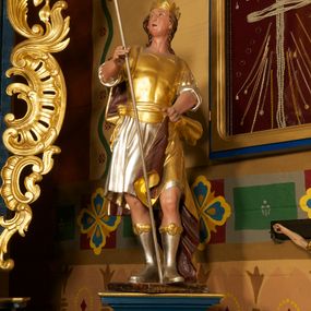 Zdjęcie nr 1: Figura ustawiona na niskiej, prostokątnej podstawie. Ukazana frontalnie, w wykroku lewej nogi, w lekkim odchyleniu ciała, z uniesioną głową skierowaną w lewo. Obie ręce ma wyciągnięte przed siebie, w prawej trzyma włócznię. Twarz ma szeroką, z niskim czołem i podkreślonym podbródkiem, o małych, migdałowatych oczach, niewielkim nosie i ustach. Włosy rzeźbione w lekko falowane pasma, ściśle przylegają do głowy i opadają na kark. Postać ma na głowie koronę otwartą. Ubrana jest w srebrzoną tunikę, sięgającą kolan, z podwiniętymi rękawami, złoconą zbroję tortową z piórami na ramionach, przewiązaną szarfą oraz złocony płaszcz z brązową podszewką, zarzucony na prawe ramię i opadający na lewą nogę. Na nogach ma srebrzone, wysokie buty. Polichromia ciała naturalistyczna.
