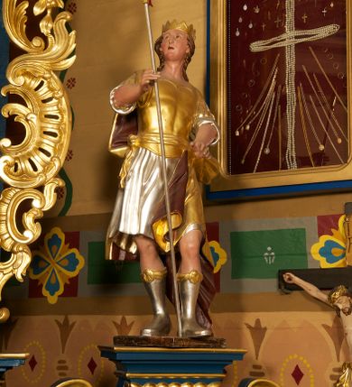 Zdjęcie nr 2: Figura ustawiona na niskiej, prostokątnej podstawie. Ukazana frontalnie, w wykroku lewej nogi, w lekkim odchyleniu ciała, z uniesioną głową skierowaną w lewo. Obie ręce ma wyciągnięte przed siebie, w prawej trzyma włócznię. Twarz ma szeroką, z niskim czołem i podkreślonym podbródkiem, o małych, migdałowatych oczach, niewielkim nosie i ustach. Włosy rzeźbione w lekko falowane pasma, ściśle przylegają do głowy i opadają na kark. Postać ma na głowie koronę otwartą. Ubrana jest w srebrzoną tunikę, sięgającą kolan, z podwiniętymi rękawami, złoconą zbroję tortową z piórami na ramionach, przewiązaną szarfą oraz złocony płaszcz z brązową podszewką, zarzucony na prawe ramię i opadający na lewą nogę. Na nogach ma srebrzone, wysokie buty. Polichromia ciała naturalistyczna.
