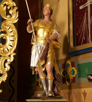 Zdjęcie nr 3: Figura ustawiona na niskiej, prostokątnej podstawie. Ukazana frontalnie, w wykroku lewej nogi, w lekkim odchyleniu ciała, z uniesioną głową skierowaną w lewo. Obie ręce ma wyciągnięte przed siebie, w prawej trzyma włócznię. Twarz ma szeroką, z niskim czołem i podkreślonym podbródkiem, o małych, migdałowatych oczach, niewielkim nosie i ustach. Włosy rzeźbione w lekko falowane pasma, ściśle przylegają do głowy i opadają na kark. Postać ma na głowie koronę otwartą. Ubrana jest w srebrzoną tunikę, sięgającą kolan, z podwiniętymi rękawami, złoconą zbroję tortową z piórami na ramionach, przewiązaną szarfą oraz złocony płaszcz z brązową podszewką, zarzucony na prawe ramię i opadający na lewą nogę. Na nogach ma srebrzone, wysokie buty. Polichromia ciała naturalistyczna.
