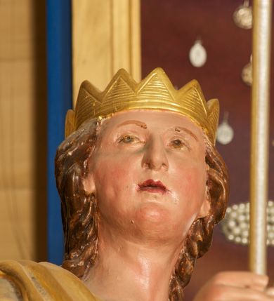 Zdjęcie nr 4: Figura ustawiona na niskiej, prostokątnej podstawie. Ukazana frontalnie, w wykroku lewej nogi, w lekkim odchyleniu ciała, z uniesioną głową skierowaną w lewo. Obie ręce ma wyciągnięte przed siebie, w prawej trzyma włócznię. Twarz ma szeroką, z niskim czołem i podkreślonym podbródkiem, o małych, migdałowatych oczach, niewielkim nosie i ustach. Włosy rzeźbione w lekko falowane pasma, ściśle przylegają do głowy i opadają na kark. Postać ma na głowie koronę otwartą. Ubrana jest w srebrzoną tunikę, sięgającą kolan, z podwiniętymi rękawami, złoconą zbroję tortową z piórami na ramionach, przewiązaną szarfą oraz złocony płaszcz z brązową podszewką, zarzucony na prawe ramię i opadający na lewą nogę. Na nogach ma srebrzone, wysokie buty. Polichromia ciała naturalistyczna.
