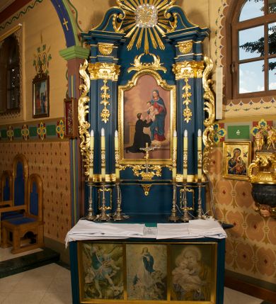 Zdjęcie nr 8: Obraz z przedstawieniem św. Stanisława Kostki z Matką Boską i Dzieciątkiem ujęty szeroką, złoconą ramą zamkniętą od góry łukiem nadwieszonym z uskokiem. Kompozycja skośna, po prawej stronie na obłokach stoi Matka Boska zwrócona delikatnie w prawo, która trzyma przed świętym na wyciągniętych rękach Dzieciątko Jezus. Maria ma owalną twarz, wysokie czoło, przymknięte oczy, wąski nos i małe usta;  jej głowę otacza wieniec z 12 gwiazd. Jest ubrana w czerwoną suknię i niebieski płaszcz z liliową podszewką, który niemal całkiem zakrywa jej jasne włosy. Płaszcz jest spięty na piersi i zarzucony na ramiona oraz nogi Marii. Trzymane przez nią Dzieciątko jest przepasane białą tkaniną, a obie ręce wyciąga w stronę świętego. Ma okrągłą twarz, krótkie jasne włosy, małe oczy, prosty nos i niewielkie usta; jego głowę otaczają złote promienie. Przed nimi na lewym kolanie klęczy św. Stanisław Kostka ukazany w trzech czwartych od tyłu, zwrócony w prawo. Obie ręce wyciąga w stronę Dzieciątka. Ma młodzieńczą twarz, krótkie, ciemne włosy. Ubrany jest w czarny strój zakonny, na nogach ma czarne buty. Przed nim, na stopniu gałązka lilii leży na otwartej księdze, za nim widoczny jest fragment pulpitu. Scena ukazana we wnętrzu, wypełnionym jasnofioletowymi i fioletowo-żółtymi obłokami; widoczny u dołu obrazu fragment podłogi w kolorze jasnobrązowym.