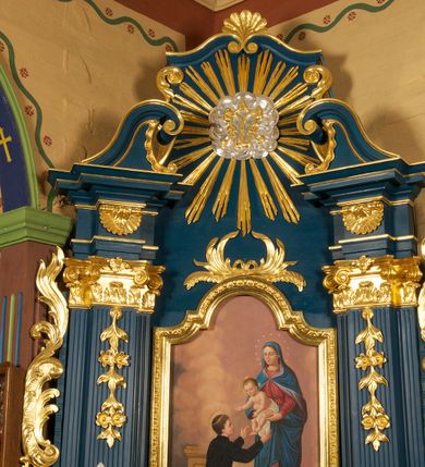 Zdjęcie nr 3: Obraz z przedstawieniem św. Stanisława Kostki z Matką Boską i Dzieciątkiem ujęty szeroką, złoconą ramą zamkniętą od góry łukiem nadwieszonym z uskokiem. Kompozycja skośna, po prawej stronie na obłokach stoi Matka Boska zwrócona delikatnie w prawo, która trzyma przed świętym na wyciągniętych rękach Dzieciątko Jezus. Maria ma owalną twarz, wysokie czoło, przymknięte oczy, wąski nos i małe usta;  jej głowę otacza wieniec z 12 gwiazd. Jest ubrana w czerwoną suknię i niebieski płaszcz z liliową podszewką, który niemal całkiem zakrywa jej jasne włosy. Płaszcz jest spięty na piersi i zarzucony na ramiona oraz nogi Marii. Trzymane przez nią Dzieciątko jest przepasane białą tkaniną, a obie ręce wyciąga w stronę świętego. Ma okrągłą twarz, krótkie jasne włosy, małe oczy, prosty nos i niewielkie usta; jego głowę otaczają złote promienie. Przed nimi na lewym kolanie klęczy św. Stanisław Kostka ukazany w trzech czwartych od tyłu, zwrócony w prawo. Obie ręce wyciąga w stronę Dzieciątka. Ma młodzieńczą twarz, krótkie, ciemne włosy. Ubrany jest w czarny strój zakonny, na nogach ma czarne buty. Przed nim, na stopniu gałązka lilii leży na otwartej księdze, za nim widoczny jest fragment pulpitu. Scena ukazana we wnętrzu, wypełnionym jasnofioletowymi i fioletowo-żółtymi obłokami; widoczny u dołu obrazu fragment podłogi w kolorze jasnobrązowym.