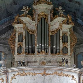 Zdjęcie nr 1: Szafa organowa architektoniczna, pięcioosiowa, na podwójnym cokole, o wklęsło-wypukłym licu. Osie wydzielone lizenami. Środkowa na planie wypukłym, najwyższa, zamknięta niepełnym belkowaniem, z gzymsem przybierającym kształt łuku segmentowego nadwieszonego oraz prześwitem na piszczałki w formie stojącego prostokąta. Dwie osie wewnętrzne wklęsłe, niższe, zamknięte niepełnym belkowaniem przyjmującym esowaty zarys; z prześwitami na piszczałki w formie zbliżonej do stojącego prostokąta o wklęsłym górnym boku. Dwie osie zewnętrzne najniższe, zamknięte niepełnym belkowaniem z podwójnymi prześwitami na piszczałki w formie stojącego prostokąta. Cokół o fantazyjnym, wklęsło-wypukłym zarysie, dekorowany pośrodku muszlą. Szafa dekorowana ażurowym rocaille'm w partii prześwitów na piszczałki, lizen, uszu, szczytów na belkowaniu. Ponadto na dekoracji gzymsu siedzą cztery muzykujące putta. Szafa organowa w całości polichromowana na sposób imitujący marmoryzację w kolorze białym z szarym żyłkowaniem, ornamentyka złocona.