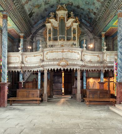 Zdjęcie nr 2: Szafa organowa architektoniczna, pięcioosiowa, na podwójnym cokole, o wklęsło-wypukłym licu. Osie wydzielone lizenami. Środkowa na planie wypukłym, najwyższa, zamknięta niepełnym belkowaniem, z gzymsem przybierającym kształt łuku segmentowego nadwieszonego oraz prześwitem na piszczałki w formie stojącego prostokąta. Dwie osie wewnętrzne wklęsłe, niższe, zamknięte niepełnym belkowaniem przyjmującym esowaty zarys; z prześwitami na piszczałki w formie zbliżonej do stojącego prostokąta o wklęsłym górnym boku. Dwie osie zewnętrzne najniższe, zamknięte niepełnym belkowaniem z podwójnymi prześwitami na piszczałki w formie stojącego prostokąta. Cokół o fantazyjnym, wklęsło-wypukłym zarysie, dekorowany pośrodku muszlą. Szafa dekorowana ażurowym rocaille'm w partii prześwitów na piszczałki, lizen, uszu, szczytów na belkowaniu. Ponadto na dekoracji gzymsu siedzą cztery muzykujące putta. Szafa organowa w całości polichromowana na sposób imitujący marmoryzację w kolorze białym z szarym żyłkowaniem, ornamentyka złocona.