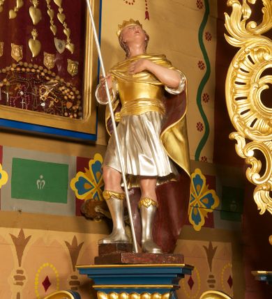 Zdjęcie nr 2: Figura ustawiona na niskiej, prostokątnej podstawie. Ukazana frontalnie, w silnym odchyleniu ciała, z uniesioną głową skierowaną w prawo. Lewą rękę kładzie na piersi, w prawej, wyciągniętej do przodu trzyma włócznię. Lewa noga ugięta, oparta na stopniu. Twarz ma szeroką, z niskim czołem i podkreślonym podbródkiem, o małych, migdałowatych oczach, niewielkim nosie i ustach. Włosy rzeźbione w lekko falowane pasma, ściśle przylegają do głowy i opadają na kark. Postać ma na głowie koronę otwartą. Ubrana jest w srebrzoną tunikę, sięgającą kolan, z podwiniętymi rękawami, złocony napierśnik przewiązany szarfą oraz złocony płaszcz z brązową podszewką, zarzucony pod szyją i opadający na plecy. Na nogach ma srebrzone, wysokie buty. Polichromią ciała naturalistyczna. 



