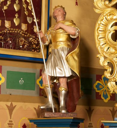 Zdjęcie nr 3: Figura ustawiona na niskiej, prostokątnej podstawie. Ukazana frontalnie, w silnym odchyleniu ciała, z uniesioną głową skierowaną w prawo. Lewą rękę kładzie na piersi, w prawej, wyciągniętej do przodu trzyma włócznię. Lewa noga ugięta, oparta na stopniu. Twarz ma szeroką, z niskim czołem i podkreślonym podbródkiem, o małych, migdałowatych oczach, niewielkim nosie i ustach. Włosy rzeźbione w lekko falowane pasma, ściśle przylegają do głowy i opadają na kark. Postać ma na głowie koronę otwartą. Ubrana jest w srebrzoną tunikę, sięgającą kolan, z podwiniętymi rękawami, złocony napierśnik przewiązany szarfą oraz złocony płaszcz z brązową podszewką, zarzucony pod szyją i opadający na plecy. Na nogach ma srebrzone, wysokie buty. Polichromią ciała naturalistyczna. 



