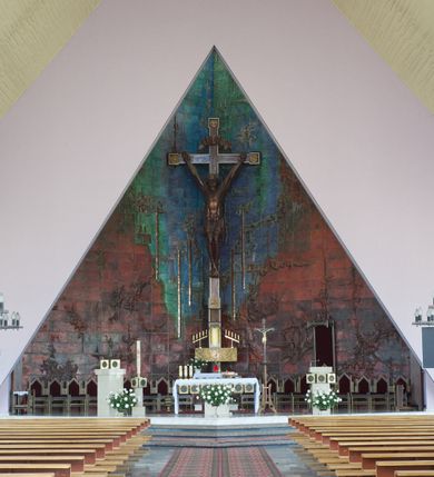 Zdjęcie nr 1: Okładzina ceramiczna pokrywa dwie ściany zamykające prezbiterium, łączące się pod kątem rozwartym i doświetlone pośrodku przez wpadające z górnego świetlika światło. Płyty są płaskorzeźbione i opracowane reliefowo, w kolorystyce błękitnej i seledynowej spływającej od szczytu ściany i przechodzącej w intensywną czerwień i brązy.