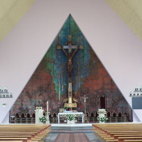 Zdjęcie nr 1: Okładzina ceramiczna pokrywa dwie ściany zamykające prezbiterium, łączące się pod kątem rozwartym i doświetlone pośrodku przez wpadające z górnego świetlika światło. Płyty są płaskorzeźbione i opracowane reliefowo, w kolorystyce błękitnej i seledynowej spływającej od szczytu ściany i przechodzącej w intensywną czerwień i brązy.